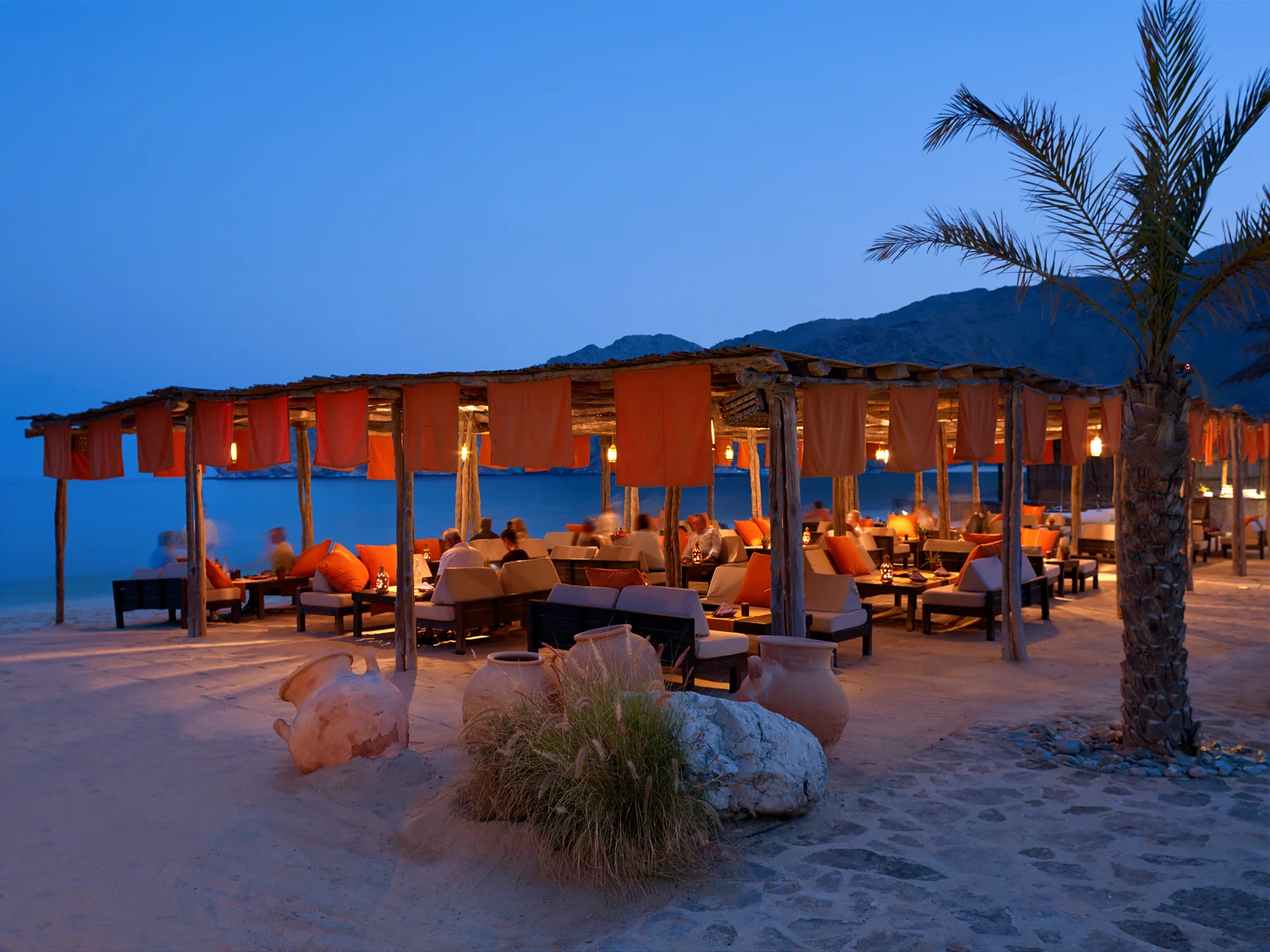 Shua Shack on the beach at blue hour, an open timber frame hung with orange fabric panels and lanterns, guests seated at low tables on the sand with large clay jars and a palm in the foreground.