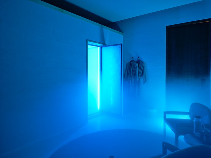 Soft blue light streams in from an open door, illuminating two robes on the wall, a nearby chair, and gentle mist for a tranquil spa-inspired setting.
