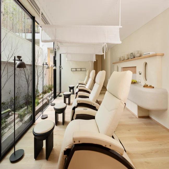 Reclining chairs with footstools in a light-filled spa space, large windows open to a green garden, inviting relaxation and connection with nature.