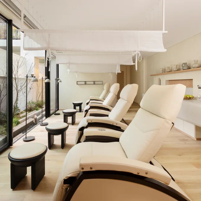 Reclining chairs with footstools in a light-filled spa space, large windows open to a green garden, inviting relaxation and connection with nature.