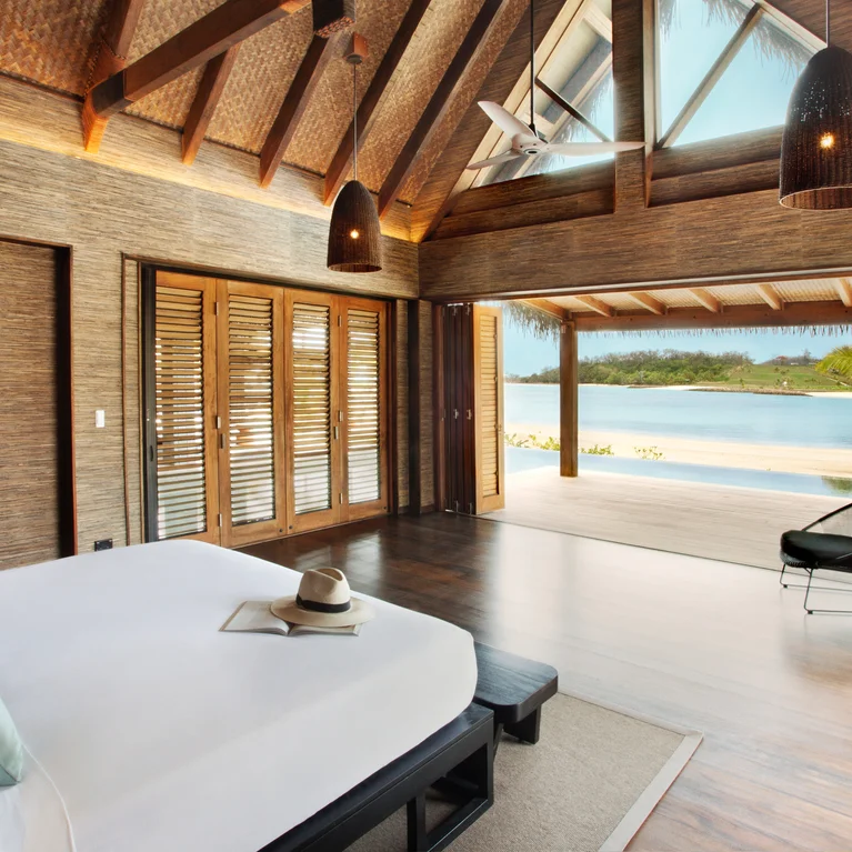 带有天然木质细节的卧室通向可俯瞰海滩和海洋的露台。图为床上的床、椅子和太阳帽。.