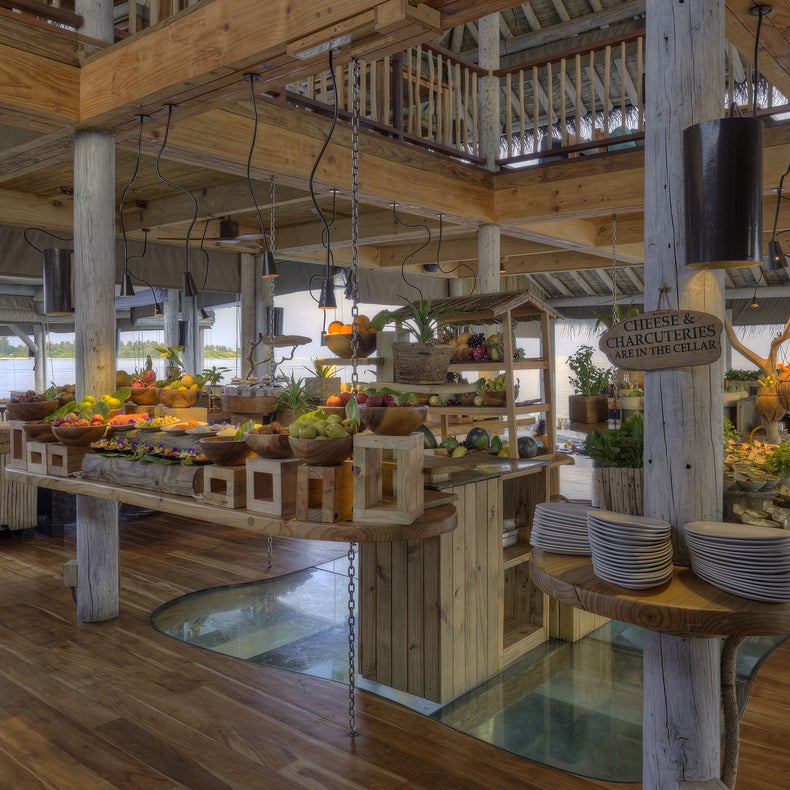 An open-air buffet features wooden beams and tables with a selection of fruits, cheeses, and dishes. Plates and utensils are arranged at one end. Large windows offer an ocean view.