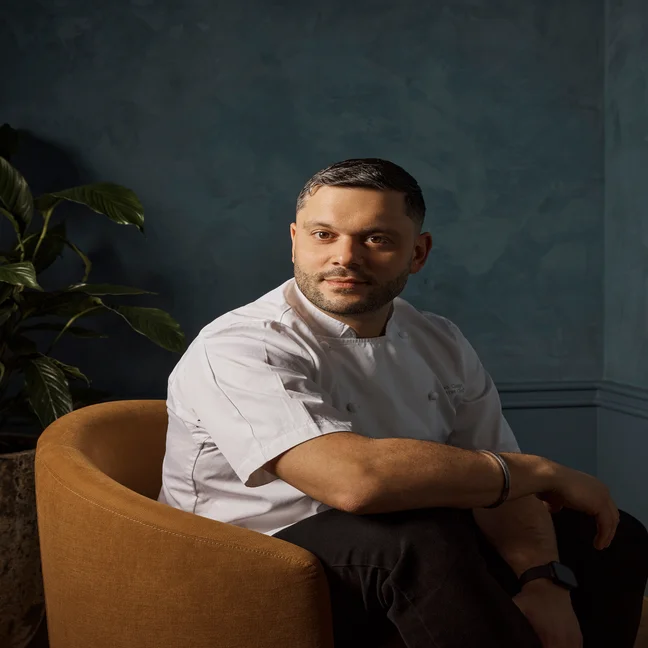 A chef in a white coat sits in a brown chair near a dark blue wall, gazing calmly to the side. A vibrant green plant adds natural texture to the background.