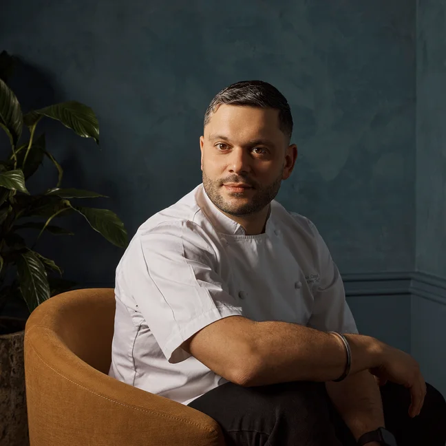 A chef in a white coat sits in a brown chair near a dark blue wall, gazing calmly to the side. A vibrant green plant adds natural texture to the background.
