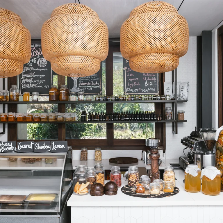 Café interior featuring wicker pendant lights, glass jars of honey and dried fruits, chalkboard menus, and a dessert counter beside large windows.