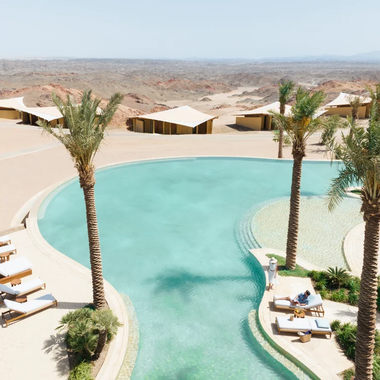 A desert retreat featuring palm trees, a curved pool, lounge chairs, and canvas tents against a backdrop of rocky landscapes and open skies.