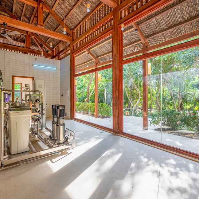 Sunlit room with floor-to-ceiling windows, indoor equipment on the left, and outdoor garden views that blend nature with thoughtful interior design.