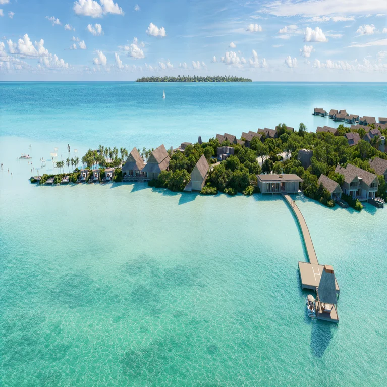 Resort villas positioned above clear turquoise water, connected by wooden docks and surrounded by trees, with another island visible across the sea beneath a bright sky.
