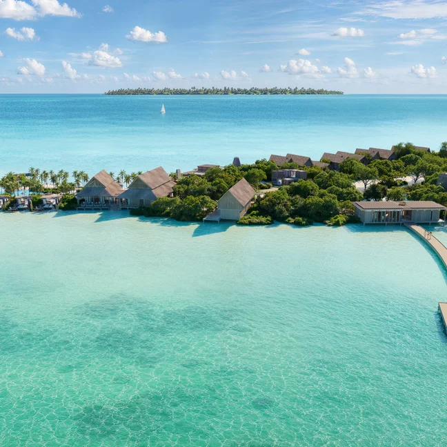 Villas de un complejo turístico situadas sobre aguas cristalinas de color turquesa, unidas por embarcaderos de madera y rodeadas de árboles, con otra isla visible al otro lado del mar bajo un cielo radiante.