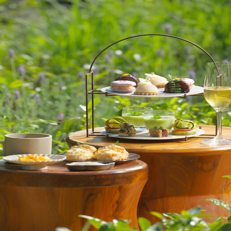 Afternoon tea with pastries, desserts, and beverages arranged on wooden tables set beside a vibrant green garden, capturing a sense of tranquility and care.