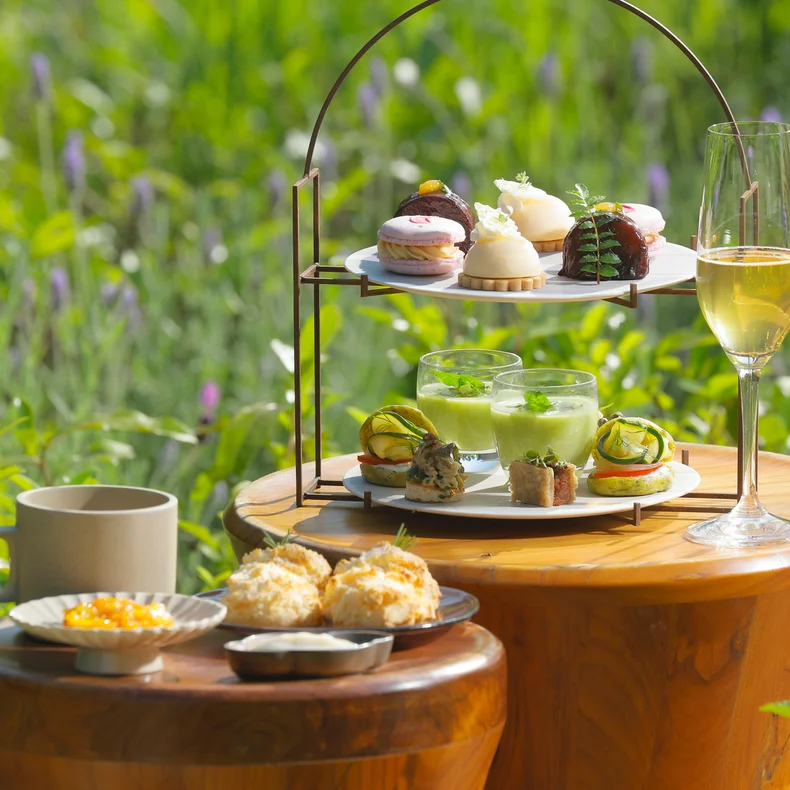 Afternoon tea with pastries, desserts, and beverages arranged on wooden tables set beside a vibrant green garden, capturing a sense of tranquility and care.