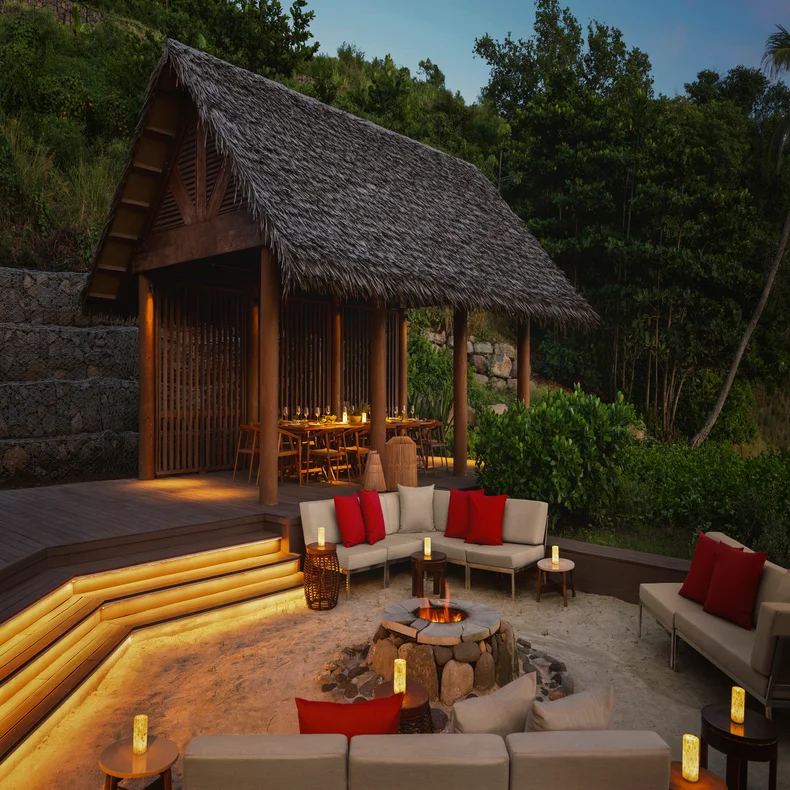 Open-air pavilion with a dining table and adjacent firepit, surrounded by cushioned seating and lanterns amid greenery at sunset.