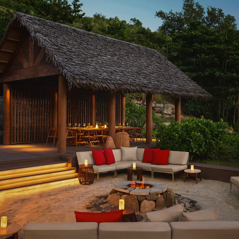 Open-air pavilion with a dining table and adjacent firepit, surrounded by cushioned seating and lanterns amid greenery at sunset.