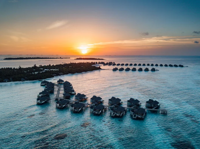 The water bungalows and villas are situated above clear turquoise waters, with islands in the distance and vibrant skies at dusk. Sustainable design in harmony with nature....