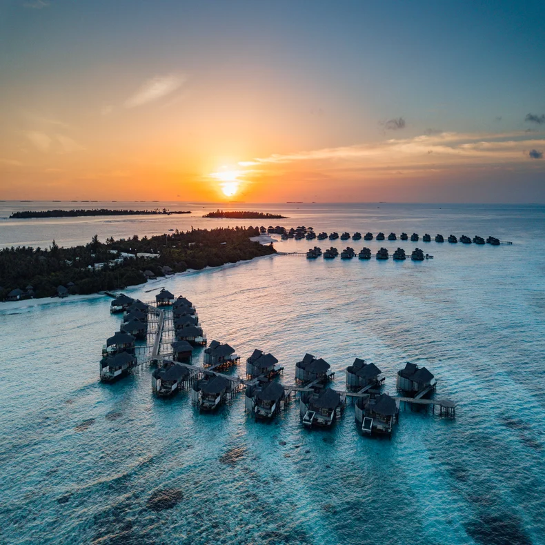 The water bungalows and villas are situated above clear turquoise waters, with islands in the distance and vibrant skies at dusk. Sustainable design in harmony with nature....