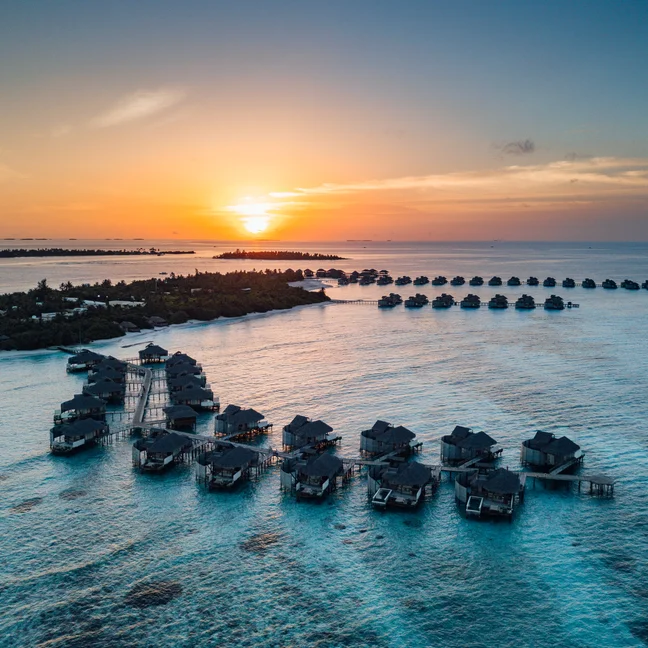 The water bungalows and villas are situated above clear turquoise waters, with islands in the distance and vibrant skies at dusk. Sustainable design in harmony with nature....