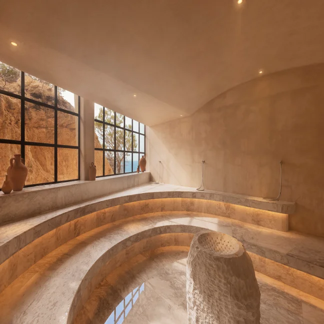 Steam room with gentle natural light, curved marble benches, clay pots, expansive windows and a central stone fountain. Designed for relaxation and wellbeing.