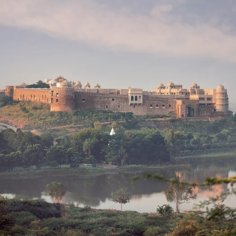歴史的な砦が緑豊かな丘の上に建ち、その前には静かな川が流れ、背景の空は穏やかな霧に包まれている。.