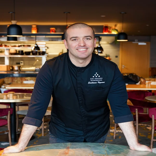 A chef in a dark uniform stands behind a marble table in a contemporary restaurant, featuring an open kitchen and red chairs.