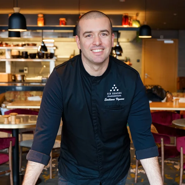 A chef in a dark uniform stands behind a marble table in a contemporary restaurant, featuring an open kitchen and red chairs.