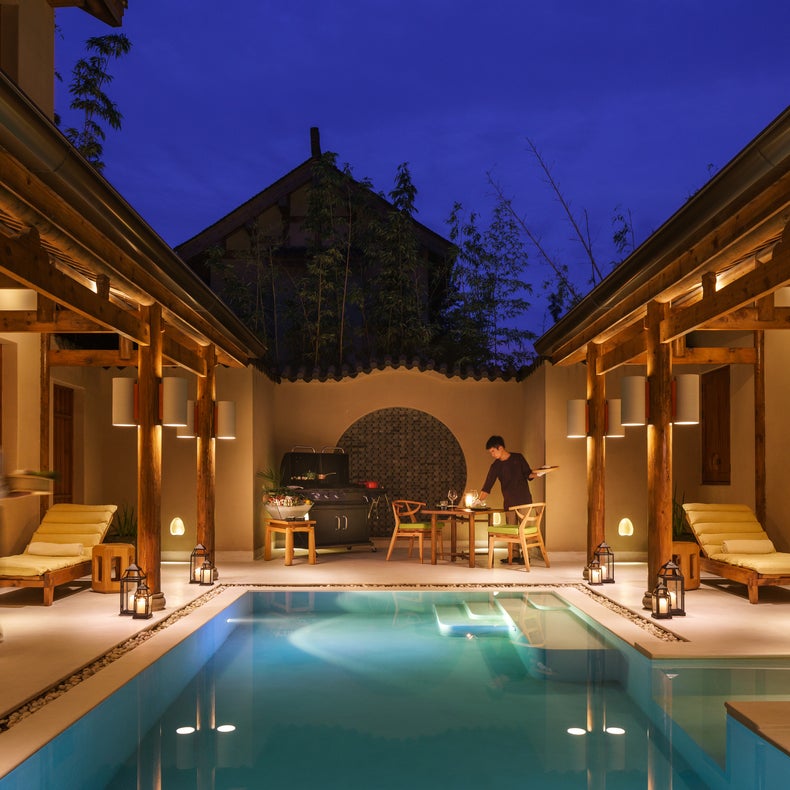 Open-air courtyard at night featuring a softly lit swimming pool, lounge chairs, two people talking at a table, and warm, gentle lighting throughout.