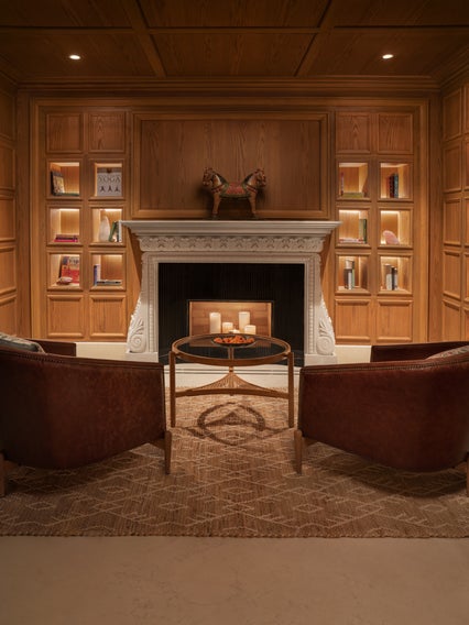 Two brown chairs face a fireplace. Candles sit on a round table. Built-in shelves hold books and decor along wood-paneled walls, creating a serene retreat.