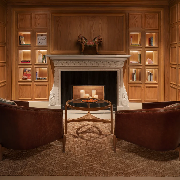 Two brown chairs face a fireplace. Candles sit on a round table. Built-in shelves hold books and decor along wood-paneled walls, creating a serene retreat.