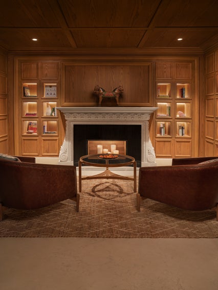 Two brown chairs face a fireplace. Candles sit on a round table. Built-in shelves hold books and decor along wood-paneled walls, creating a serene retreat.