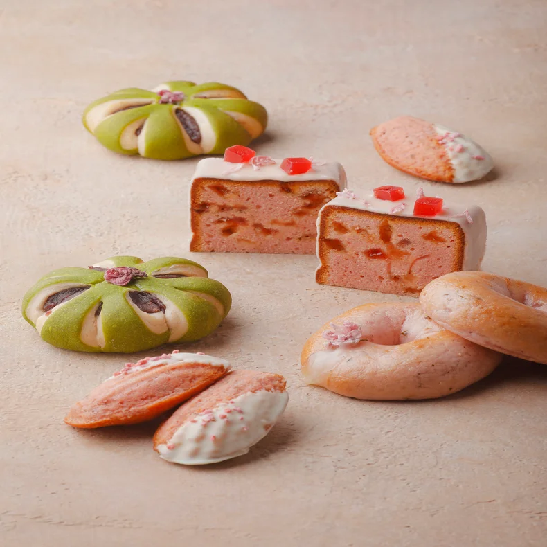 Green flower-shaped buns, pink cake slices with icing, a pink donut, and white chocolate-dipped cookies on a beige surface, presented for mindful enjoyment.