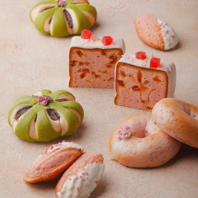 Green flower-shaped buns, pink cake slices with icing, a pink donut, and white chocolate-dipped cookies on a beige surface, presented for mindful enjoyment.