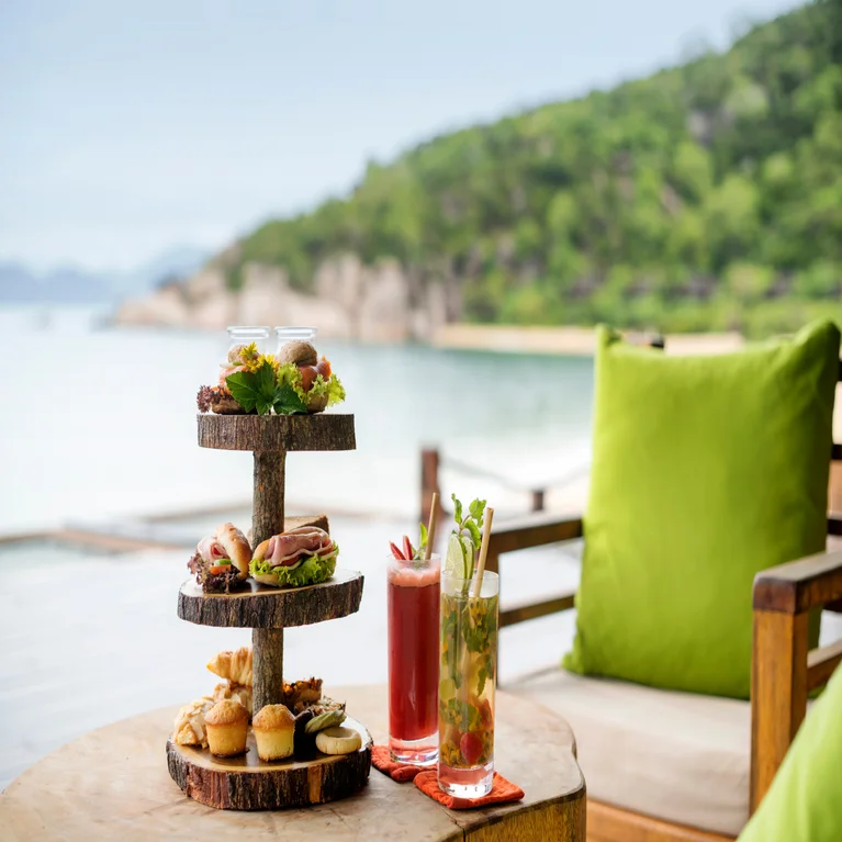 A selection of snacks on a three-level stand and two vibrant drinks set on a table by the water, with a green cushion and natural scenery in the background.
