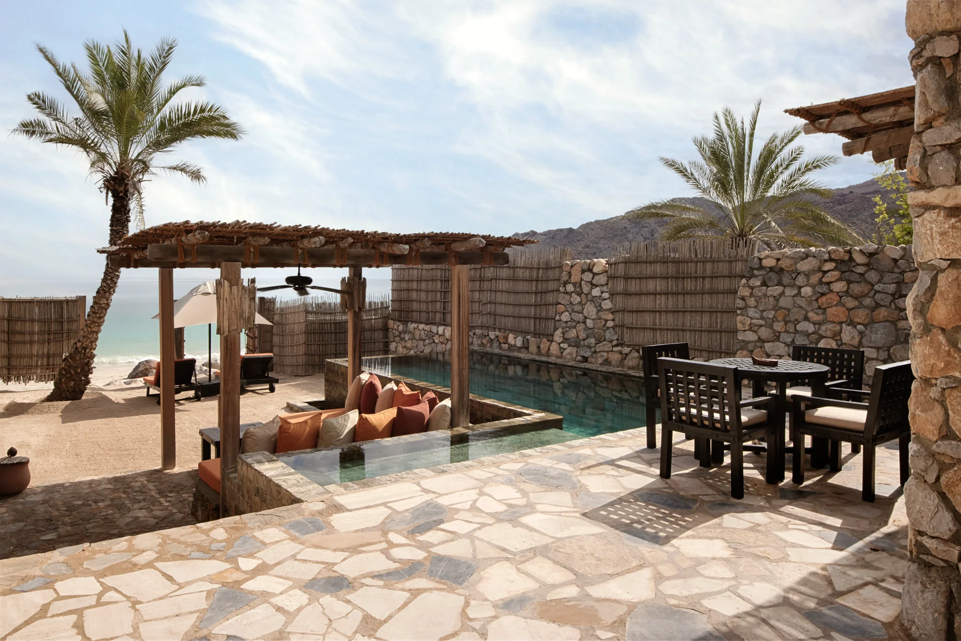A beachfront pool villa's flagstone terrace, an outdoor dining table and chairs beside a private pool, a cushioned pergola daybed alongside it and a reed screen opening onto the sand.
