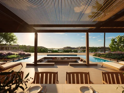 Wooden patio furniture and sun loungers beside a pool, surrounded by greenery, with views of water and the sky at dusk.