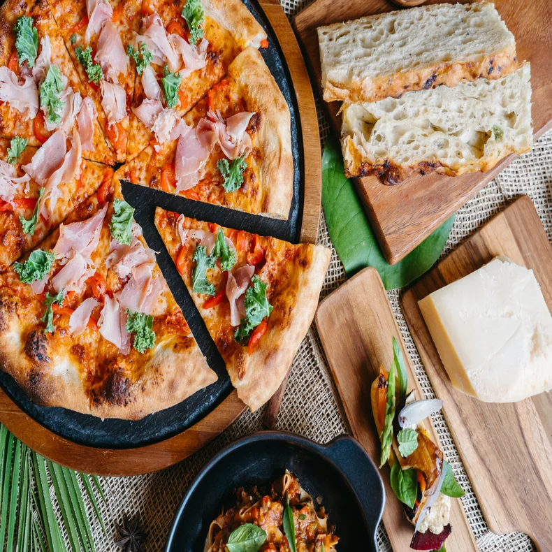 Artisan pizza topped with ham and greens, fresh sliced bread, cheese, and a small pasta bowl presented on natural wooden boards.
