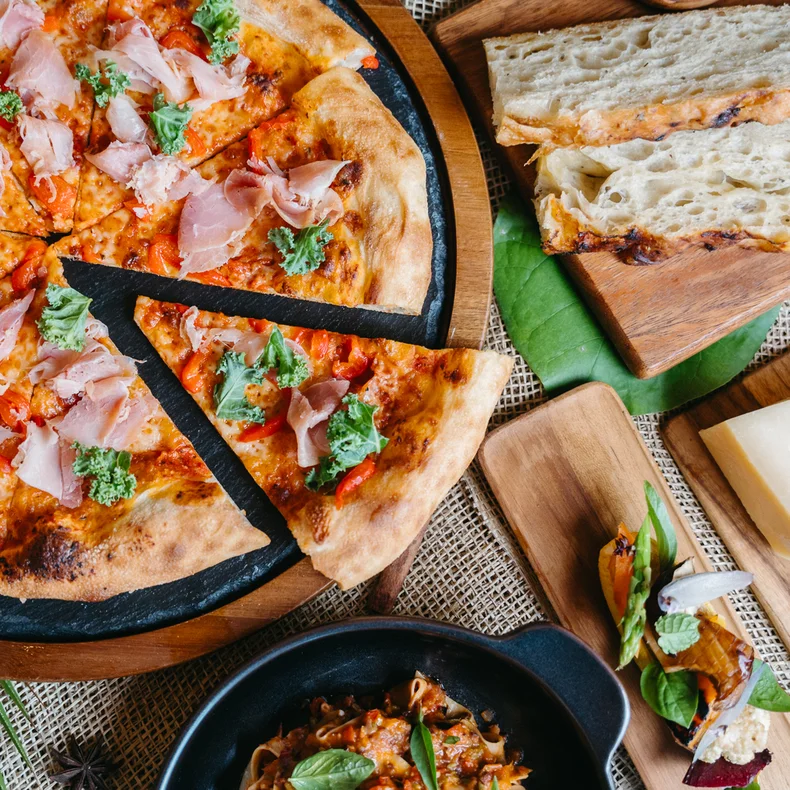 Artisan pizza topped with ham and greens, fresh sliced bread, cheese, and a small pasta bowl presented on natural wooden boards.