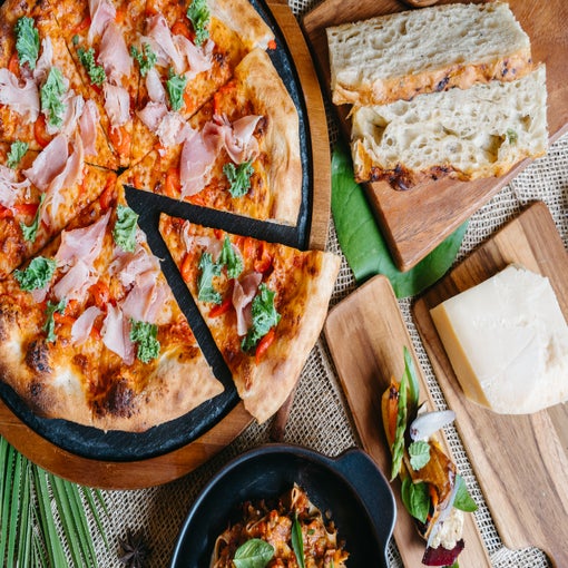Artisan pizza topped with ham and greens, fresh sliced bread, cheese, and a small pasta bowl presented on natural wooden boards.