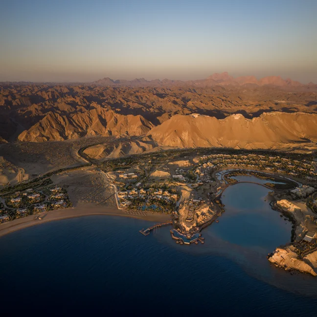 Coastal desert town bordered by mountains, sandy earth, and calm blue water at sunset, with thoughtful integration of local buildings into the natural landscape.