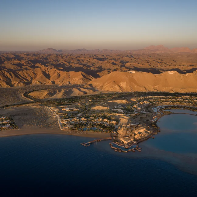 Coastal desert town bordered by mountains, sandy earth, and calm blue water at sunset, with thoughtful integration of local buildings into the natural landscape.
