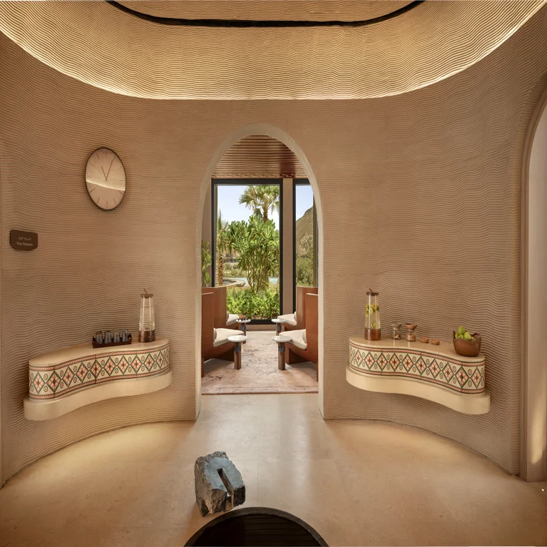 Beige space with built-in shelves, clock, drinks, and fruit. An archway leads to seating and a garden with palm trees. Daylight fills the interior for a serene atmosphere.