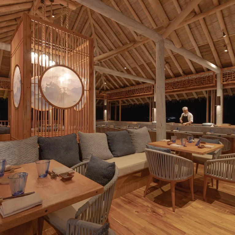 An open-air restaurant featuring wooden tables, cushioned chairs, blue glasses, and a chef preparing dishes at the counter under soft evening lighting.