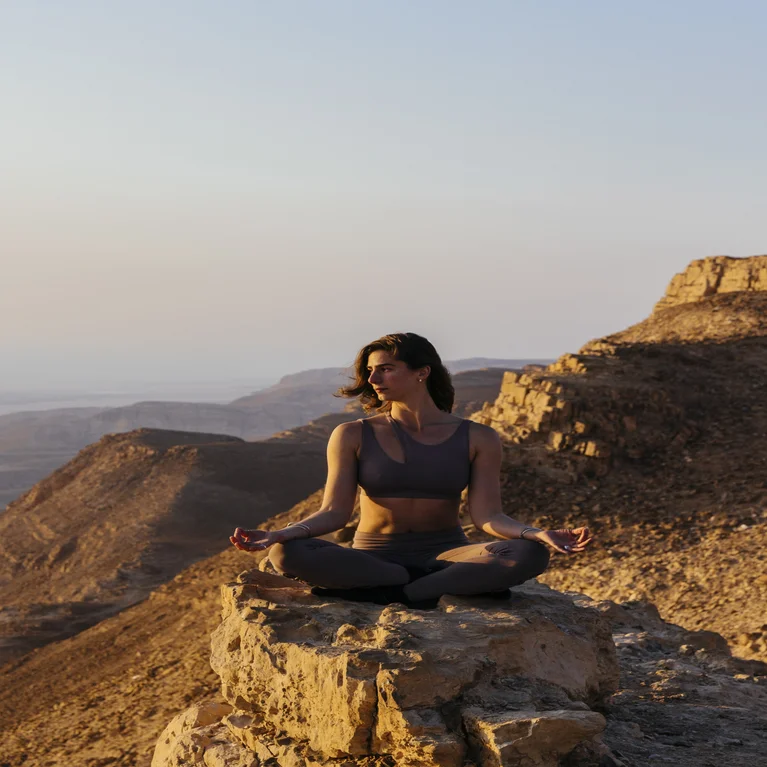 A guest in yoga clothing meditates on a rocky ledge, framed by sunlit mountains and valleys, reflecting well-being and a natural setting.