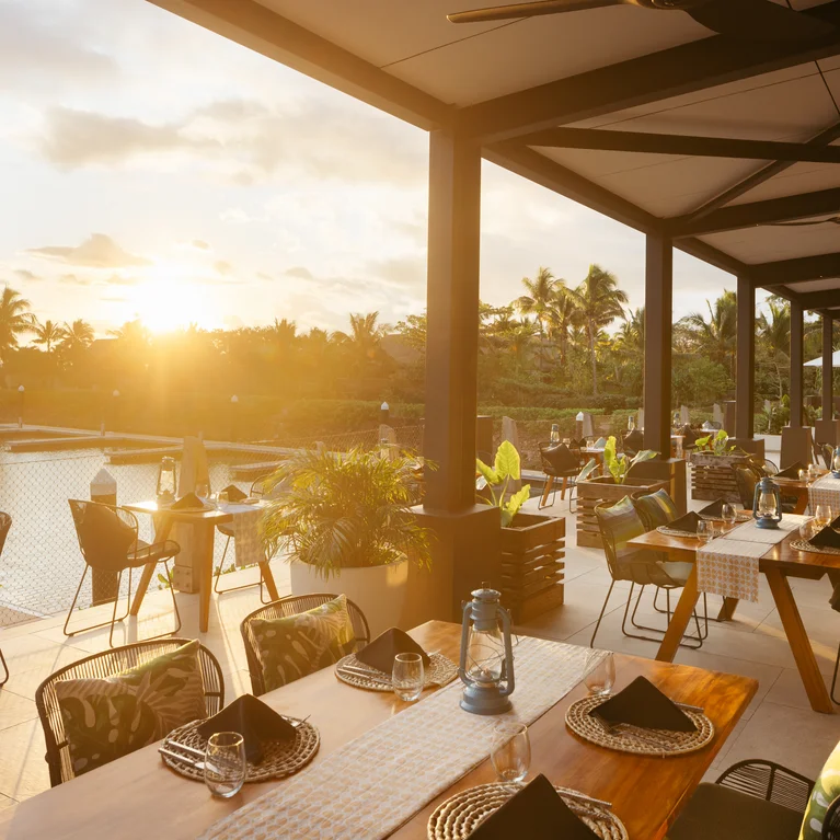 Dînez dans un patio extérieur tranquille, avec des tables disposées pour le confort, entourées de verdure tropicale et de vues sur l'eau et les palmiers au-delà.