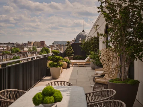 Terrace with greenery in planters, comfortable outdoor lounge seating, and cityscape visible in the background beneath a sky with scattered clouds.