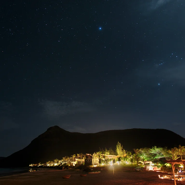 Vue nocturne des montagnes à côté d'une station balnéaire, avec des palmiers et des bâtiments d'inspiration écologique subtilement illuminés pour créer un environnement tranquille en lien avec la nature.