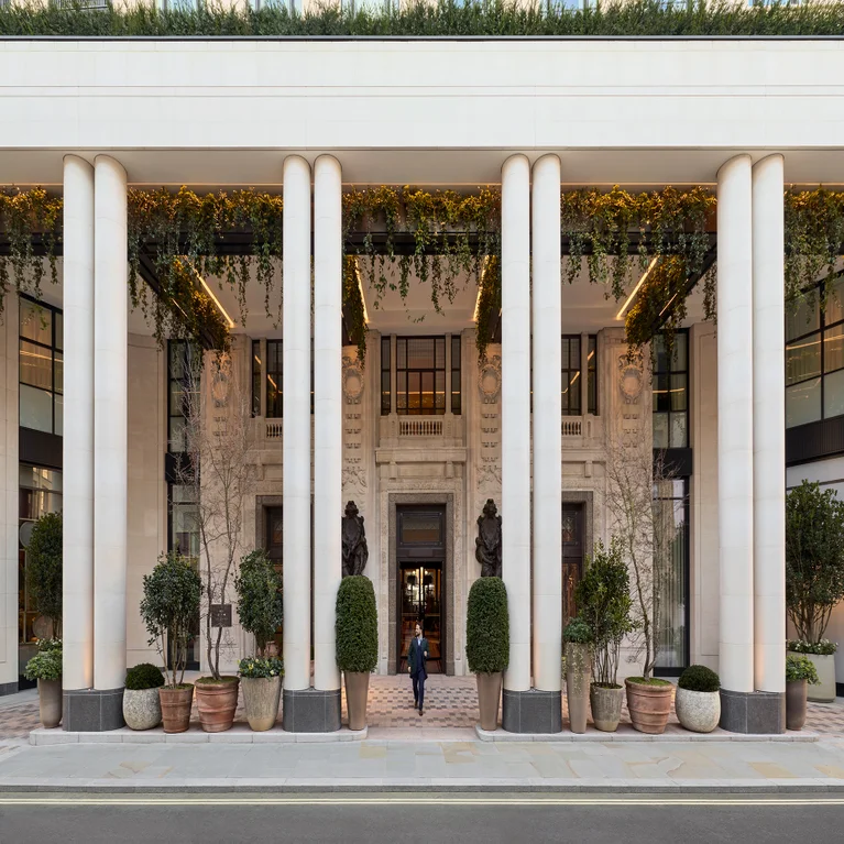 A person walks through doors framed by white columns, with potted plants and lush greenery enhancing the welcoming, nature-inspired setting.