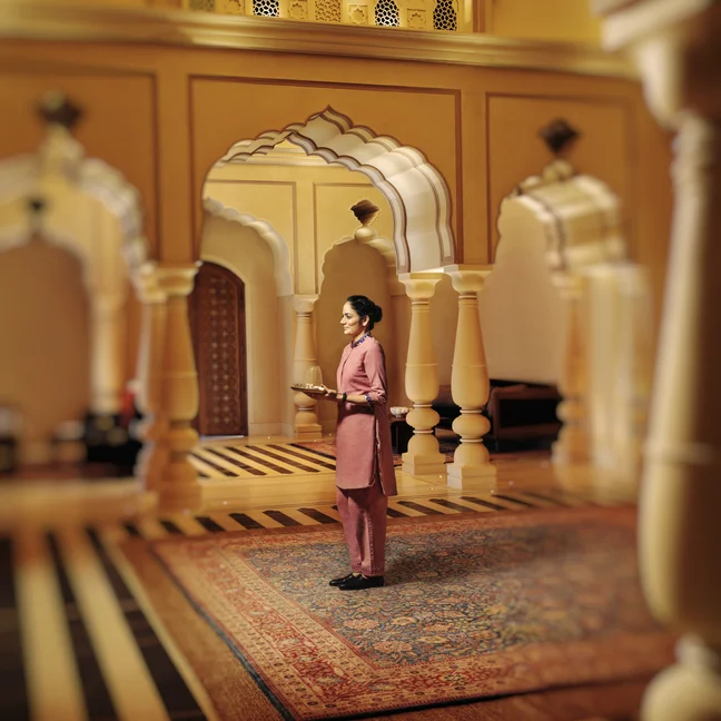 ضيف يرتدي ثياباً بنفسجية اللون ويقف على سجادة منقوشة داخل مبنى ذي أقواس وأعمدة وتفاصيل ذهبية اللون....