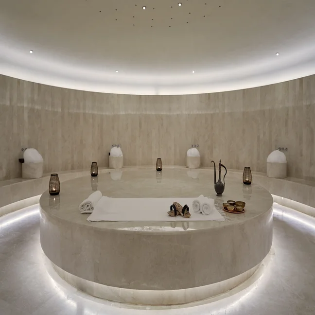 A circular marble massage platform featuring white towels, sandals, and metal bowls inside a spa room with stone walls and floor lanterns.