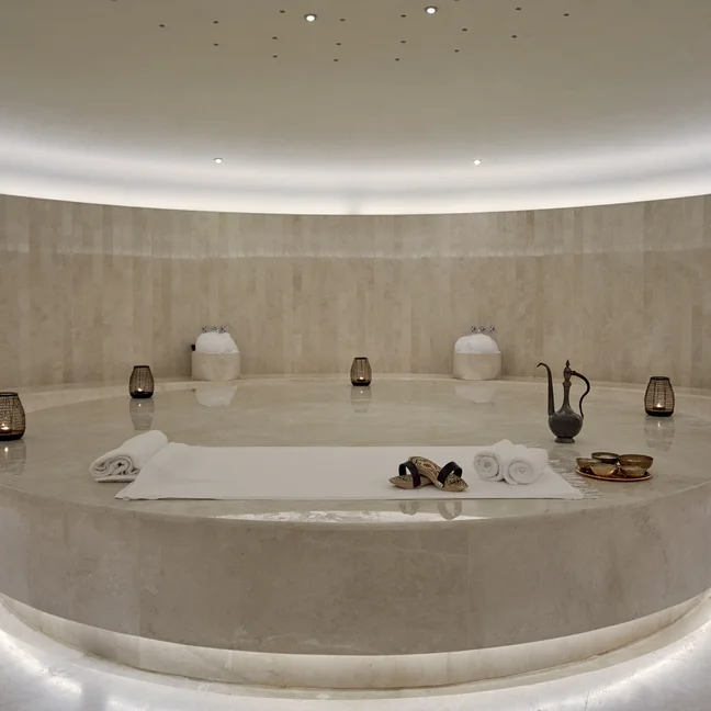 A circular marble massage platform featuring white towels, sandals, and metal bowls inside a spa room with stone walls and floor lanterns.