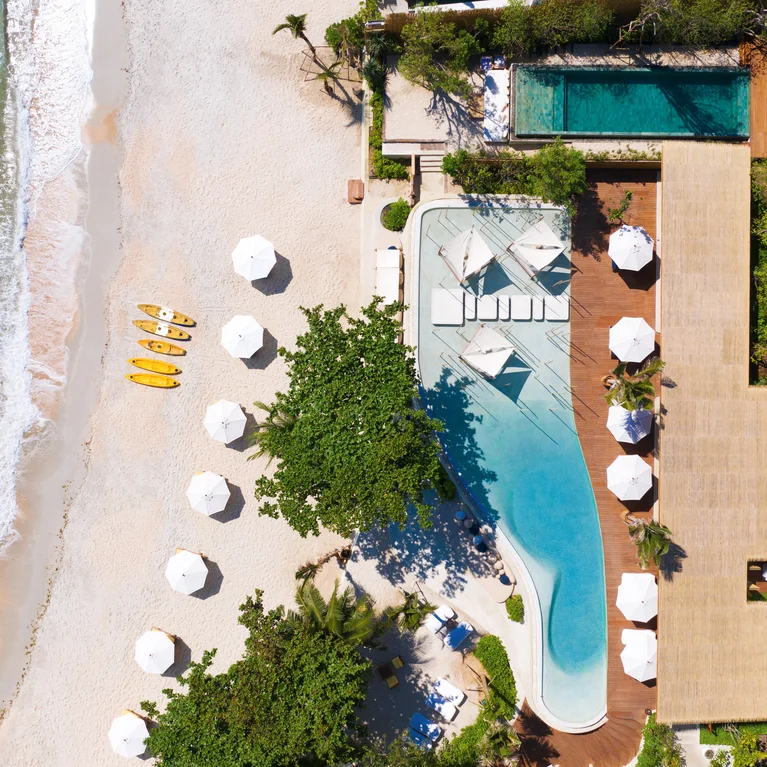 منظر لحوض سباحة المنتجع محاط بمظلات بيضاء وكراسي استرخاء بيضاء مع قوارب الكاياك على الشاطئ الرملي والمياه الزرقاء الصافية التي تلتقي بالساحل.