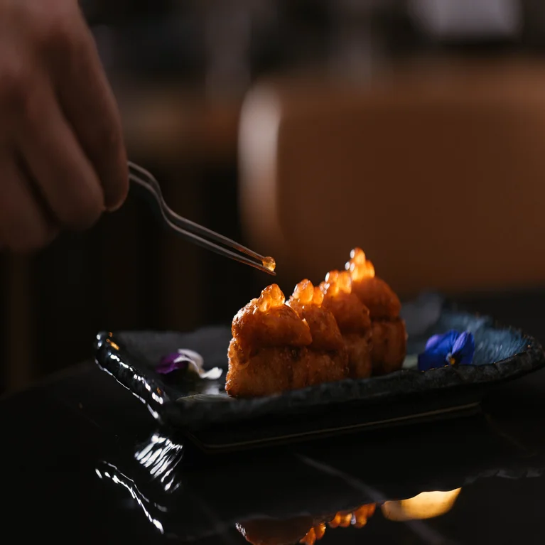 Tweezers place roe onto a bite-sized appetizer topped with uni, served on a black rectangular plate.